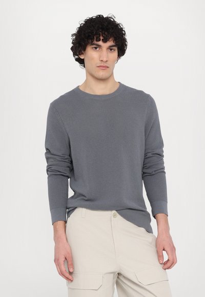 Grauer Strickpullover mit Rundhalsausschnitt und langen Ärmeln, aus strukturiertem Stoff mit taillierten Bündchen, kombiniert mit hellbeigen Cargo-Hosen.