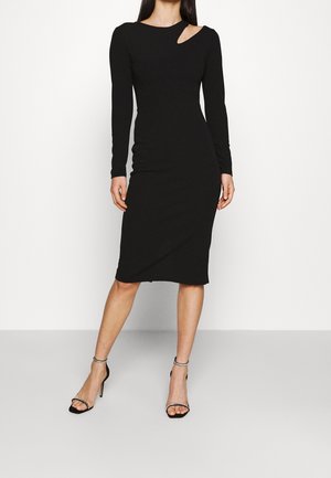 Kobieta ubrana w dopasowaną czarną sukienkę midi z długimi rękawami i małym wycięciem na ramieniu, w połączeniu z czarnymi sandałami na wysokim obcasie z paskami wokół kostek.