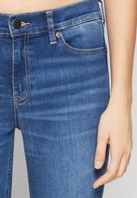Vaqueros de denim azul con una cintura de altura media, ligeras desgastadas y costuras doradas contrastantes. Presenta bolsillos frontales y un cierre de botón sencillo.