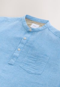 Lyseblå kortærmet skjorte med tekstureret mønster, ståkrave, tre knapper og brystlomme. Producentetiket synlig indeni.