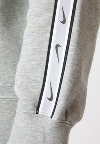 Sudadera gris con un tejido texturizado, franja lateral en blanco y negro con logotipos negros repetidos, y detalle de manga con puño.