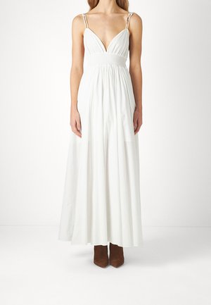 Femme portant une longue robe blanche sans manches avec un profond décolleté en V et des bottines marron, debout devant un fond clair uni.