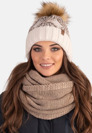 Cappello a maglia avorio con pom-pom in pelliccia marrone e dettaglio del logo, abbinato a una sciarpa infinita beige spessa e texturizzata.