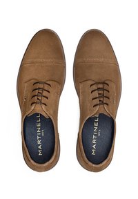 Zapatos de ante marrones con cordones, con una textura suave, punta redondeada y un interior oscuro en contraste. Presentan un sutile costura y un logo de la marca.