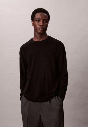 Jeune homme à la peau foncée portant une chemise noire à manches longues et un pantalon gris, debout les mains dans les poches devant un fond uni.