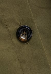 Tessuto verde oliva con un bottone nero dal design marmorizzato e quattro fori, attaccato con cuciture visibili attorno al bottone.