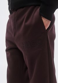 Pantaloni de trening vișinii cu textură netedă, având un logo „HUGO” subtil în relief pe coapsă și buzunare laterale.