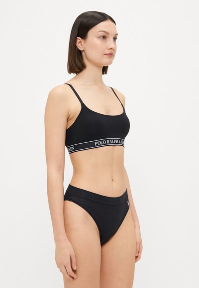 Black sports bra with thin straps and logo band; matching high-waisted bikini bottom. Smooth texture, minimalistic design, and monochrome color.