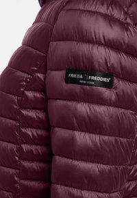 Burgundowa kurtka puchowa o pikowanej strukturze, błyszczącej powierzchni, z czarną metką z napisem "Frieda Freddies New York" na górnej części ramienia.