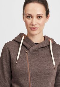 Brun zip-up hettegenser med teksturert stoff, hvite snorene og tan detaljer på hetta. Har en frontglidelås og en avslappet passform.