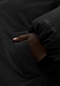 Sort jakke med en glat tekstur, hvor der er en hånd i en sidelomme. "LULULEMON" logoet er prægnet på ærmet.