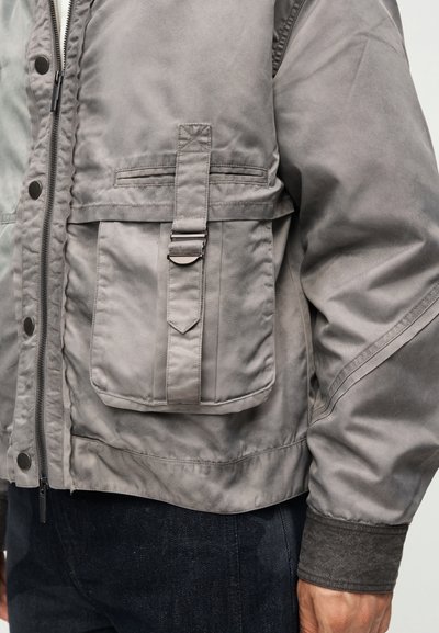 Ανοιχτόγεια μπουφάν με μπροστινό φερμουάρ, κουμπιά πατιλέτας, μεγάλη τσέπη με καπάκι, λουράκι και μεταλλική πόρπη, φορεμένο με σκούρο παντελόνι.