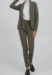 Costume rayé vert olive avec un blazer cintré et un pantalon à cordon de serrage. Tissu lisse, souligné de rayures blanches, associé à des mocassins noirs.