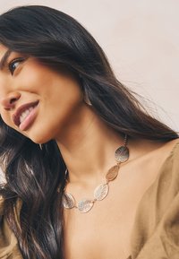 Gold-tone necklace with leaf-shaped links featuring textured patterns. The model wears matching leaf-shaped earrings and an olive green top.