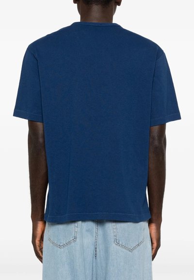 Person iført en ensfarvet blå t-shirt med korte ærmer og lyseblå denimjeans, vist bagfra mod en hvid baggrund.
