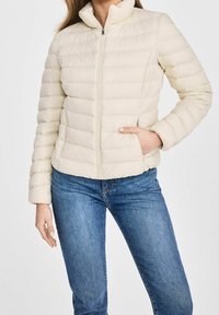 Kobieta w jasnobeżowej pikowanej kurtce puchowej z zamkiem z przodu i niebieskich dżinsach, z jedną ręką w kieszeni kurtki.
