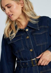 Vestido de mezclilla en azul oscuro con cuello, botones dorados y bolsillos frontales. Presenta un cinturón con una hebilla metálica rectangular y detalles de costura.