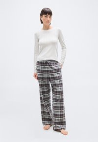 Femme portant un haut beige à manches longues et un pantalon de pyjama à carreaux gris avec de petits motifs rouges et noirs, debout pieds nus sur un fond blanc.