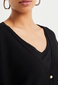 Svart ribbad cardigan med djup V-ringning, med en silverknapp på framsidan, lagrad över en svart V-ringad topp.