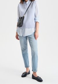 Suéter sin mangas de punto azul claro sobre una camisa azul claro, combinado con jeans azul claro y mocasines oscuros con acentos dorados.