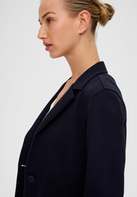 Granatowa marynarka z wyciętym kołnierzem, zapięcie na jeden guzik i wyraźnie zaznaczone ramiona. Gładki materiał o dopasowanym kroju.