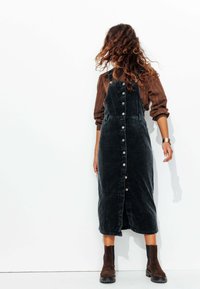 Robe salopette en velours côtelé noir avec fermeture à boutons sur le devant, poches latérales et une silhouette droite, associée à des bottes chevalières marron.