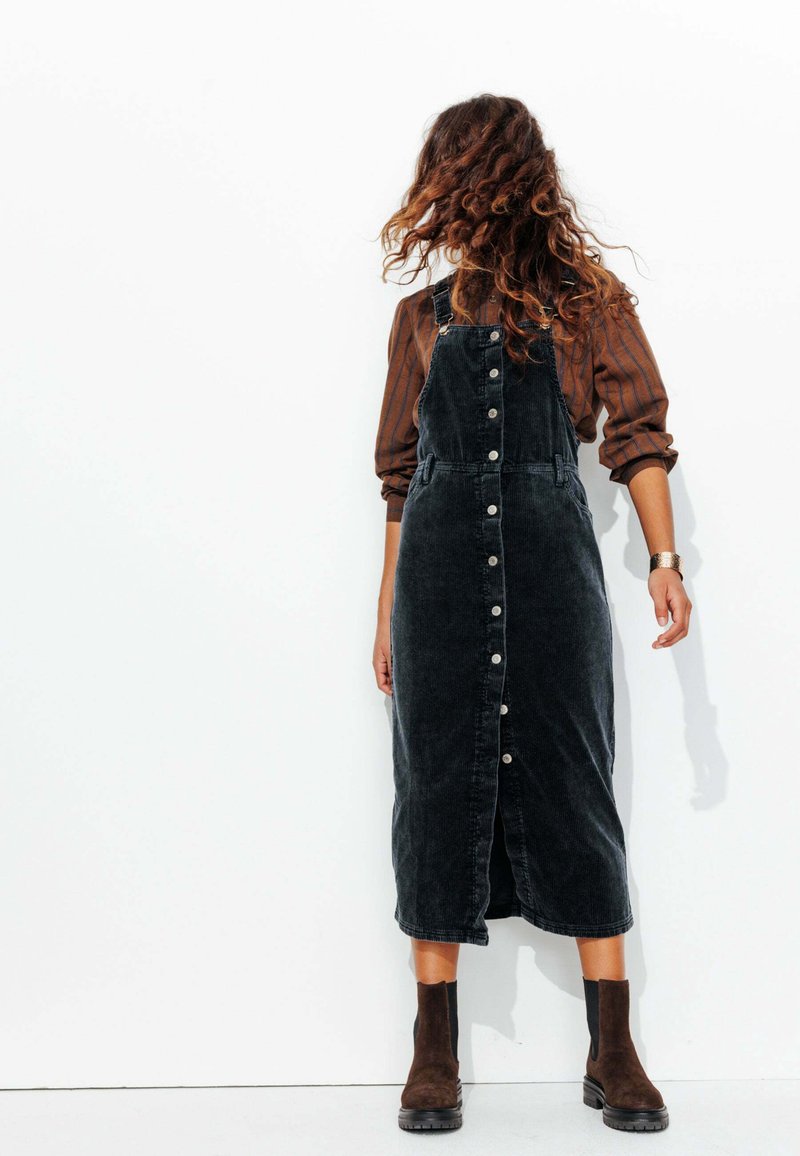 Robe salopette en velours côtelé noir avec fermeture à boutons sur le devant, poches latérales et une silhouette droite, associée à des bottes chevalières marron.