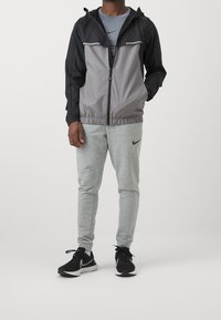 Svart och grå zipjacka med huva, elastisk midja och ljusgråa träningsoverallbyxor. Svarta sportskor med vita detaljer.