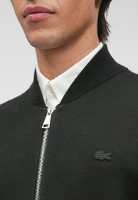 Μαύρο μπόμπερ μπουφάν από υφασμάτινο υλικό, με ραμμένο γιακά και ασημένιο φερμουάρ. Διαθέτει ένα μικρό μαύρο λογότυπο στο στήθος.