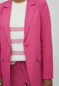 Rosa kavaj med strukturerade axlar, två knappar och sidfickor, bärs över en vit ribbad tröja med rosa ränder. Slät textur.