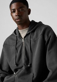 Sudadera con cremallera de color gris oscuro y textura suave, con capucha ajustable, bolsillos frontales y detalles de costura en los hombros.