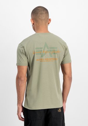 Mann mit kurzen Haaren und Ohrringen, trägt ein olivgrünes Alpha Industries T-Shirt und schwarze Hosen, zeigt den Rücken mit Logo und Schriftzug.