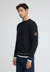 Svart kabelstickad tröja med rund hals, strukturerat m önster, vita accentränder vid ärmarna och nederkanten, med en broderad patch på ärmen.