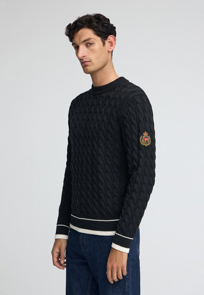 Svart kabelstickad tröja med rund hals, strukturerat mönster, vita accentränder vid ärmarna och nederkanten, med en broderad patch på ärmen.