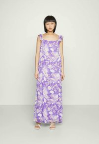 Vestido maxi lavanda com estampa floral, alças com folhos, tecido leve e um corte solto. Usado com sandálias brancas com tiras.