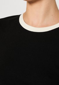 Černé žebrované tričko s kontrastním bílým kulatým výstřihem. Tkanina je hladká a má jemnou texturu, která zdůrazňuje jednoduchý a klasický design.