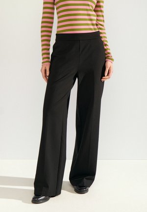 Femme portant un pantalon noir à jambes larges, des chaussures noires et un haut rayé rose et vert à manches longues, debout devant un fond uni.