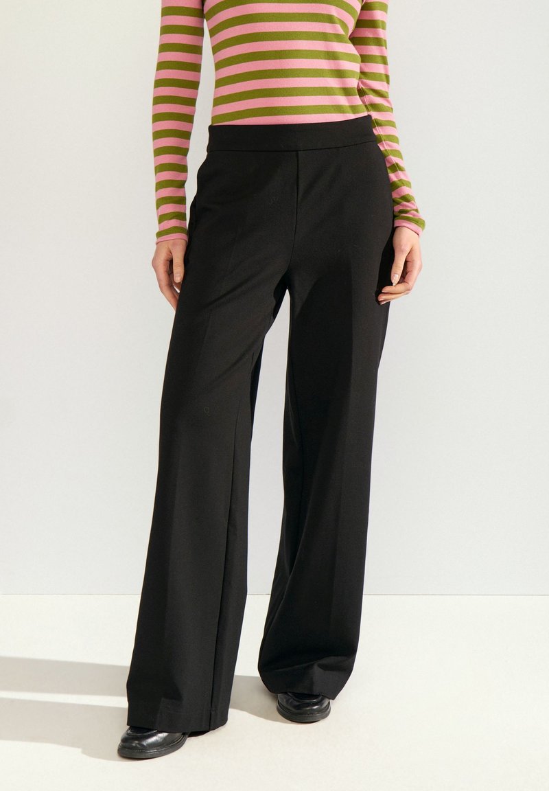 Femme portant un pantalon noir à jambes larges, des chaussures noires et un haut rayé rose et vert à manches longues, debout devant un fond uni.