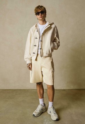 Jeune homme portant des lunettes de soleil, une veste à capuche beige, un short assorti, un t-shirt blanc, des chaussettes blanches et des baskets beiges, debout contre un mur uni.