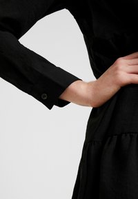 Schwarzes Hemdkleid mit tailliertem Schnitt, langen Ärmeln und geknöpften Manschetten. Der Stoff hat eine weiche Textur mit einem leichten Glanz.