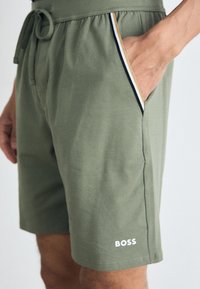 Zelené šortky vyrobené z mäkkej tkaniny, s pásom na šnurovanie, bočnými vreckami s kontrastným lemovaním a bielym logom "BOSS" na nohe.