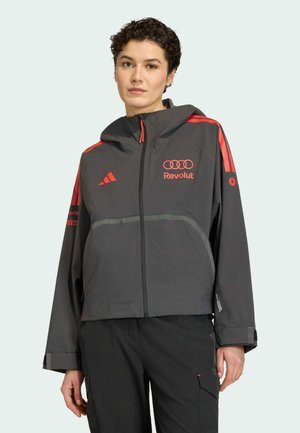 Person iført sort og rød Adidas hættetrøje med Revolut og Audi-logoer, stående mod en ensartet lys baggrund.