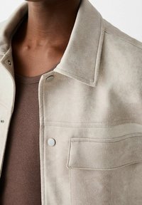 Beige mokkanahkatakki, jossa kaulus, painonapit ja etutasku. Kangas on pehmeän tuntuista ja kerroksittain ruskean topin päällä.