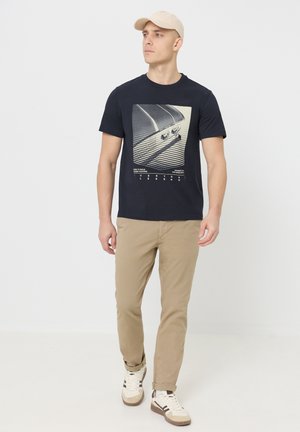 Jonge man met een beige pet, marineblauw T-shirt met print, kaki broek en witte sneakers, staand tegen een effen witte achtergrond.