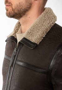 Bruine leren jack met een textuurafwerking, voorzien van een beige schapenvachtkraag en ritssluiting. Horizontale leren accenten zijn zichtbaar.