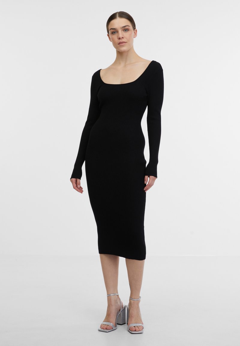 Robe noire en tricot côtelé avec décolleté carré, manches longues et silhouette ajustée, arrivant au milieu du mollet. Sandales à talons hauts en argent portées.