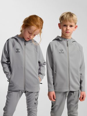 Deux enfants en vestes à capuche et pantalons gris clair assortis, l’un regarde vers le bas avec les mains dans les poches, l’autre fait face, les bras le long du corps.