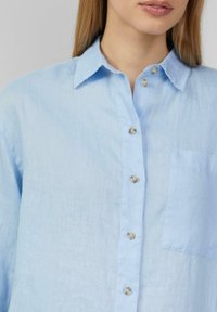 Jasnoniebieska lniana koszula z zapięciem na guziki, z ostro zakończonym kołnierzykiem, sześcioma guzikami i jedną kieszenią na piersi.