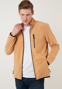Fleecejacke in beige met een hoge kraag, zwarte ritssluiting, zijzakken en zwarte accenten langs de manchetten en de zoom. Zachte textuur.