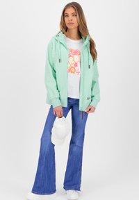 Lichtgewicht mintgroene jas met een rits, verstelbare capuchon en elastische manchetten. Gepaard met een witte t-shirt met bloemendessin en blauwe flare jeans.